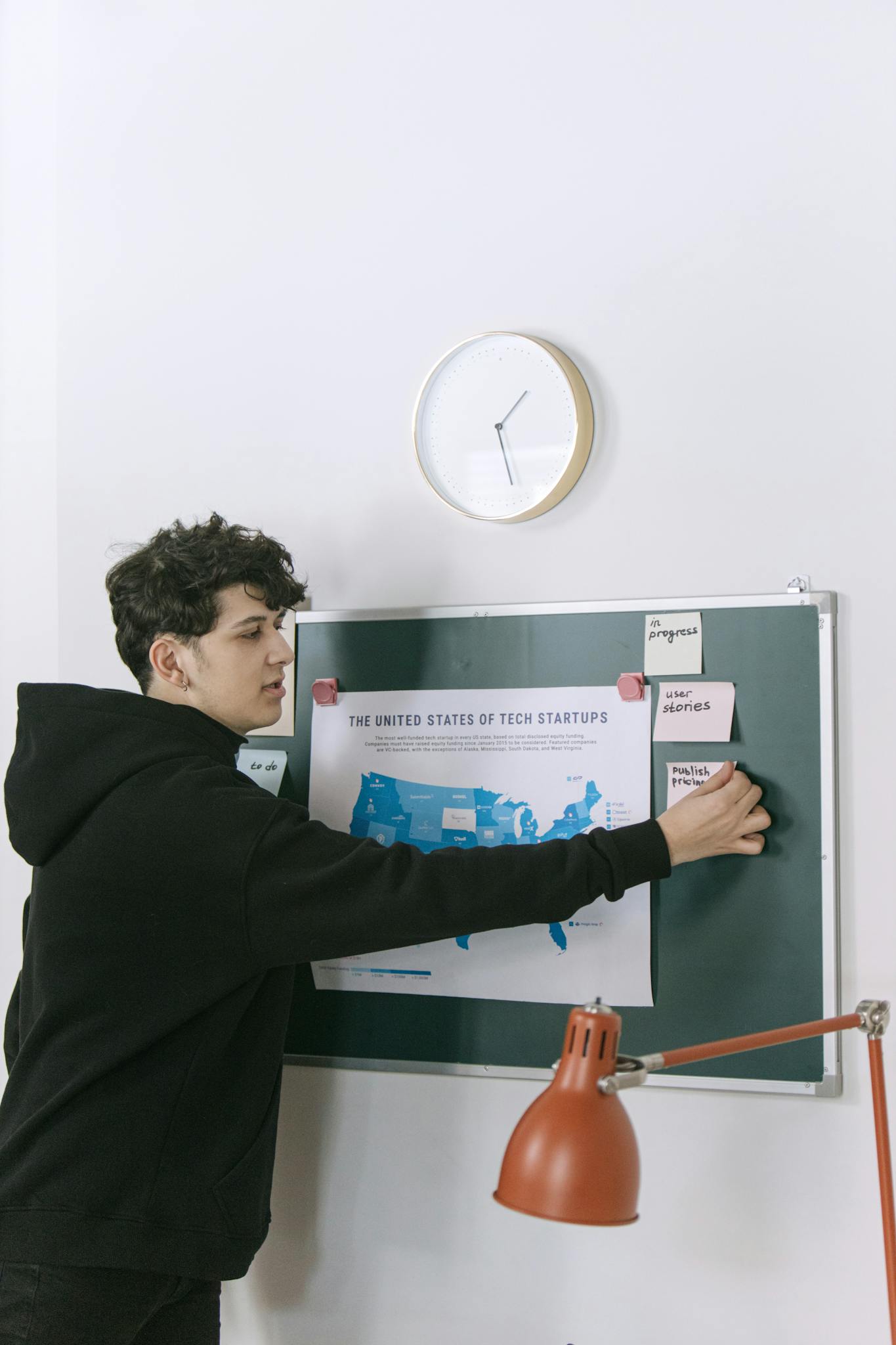 A young man in a hoodie works in a modern office setting, using a map of US tech startups for planning.
